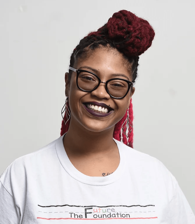 Smiling black woman in large-rimmed glasses, leaf-shapred earrings, and braided hair wears t-shirt with the Future Foundation logo